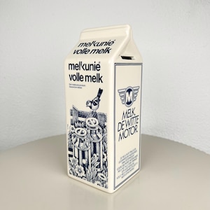 Op de afbeelding: Een crèmekleurige keramische spaarpot in de vorm van een melkpak met donkerblauwe tekst en illustraties. De voorzijde bevat de tekst "melkunie volle melk" en een afbeelding van een vogel op een hek. De zijkant toont "MELK DE WITTE MOTOR" met een M-logo.
