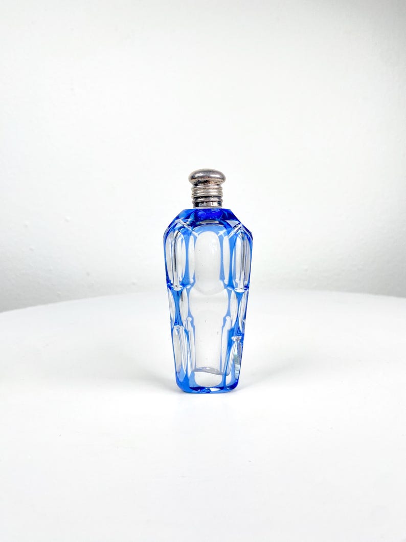 May include: A clear glass perfume bottle with blue accents and a silver-colored stopper. The bottle has a geometric design with blue lines outlining the edges and a blue rim. The bottle is empty and sits on a white surface.