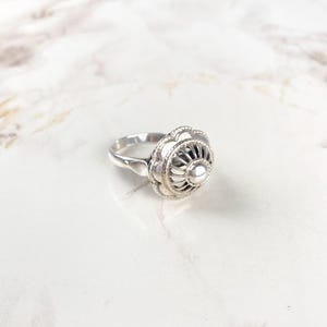 Op de afbeelding: Een zilveren ring met een bloemmotief. De ring heeft een centraal, verhoogd, cirkelvormig element omgeven door een gedetailleerd, opengewerkt patroon. De band is glad en loopt taps toe naar achteren. De ring ligt op een wit marmeren oppervlak.