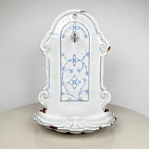 Op de afbeelding: Een witte geëmailleerde metalen wandwastafel met een geschulpte waskom en een verticaal achterpaneel versierd met blauwe bloemmotieven. Het object is voorzien van een centrale metalen kraan en vertoont roestplekken langs de randen.