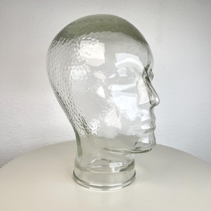 Könnte beinhalten: Ein transparenter Schaufensterkopf aus Glas mit einer texturierten, genoppten Oberfläche. Das Objekt ist in Form eines menschlichen Kopfes mit Hals gestaltet und weist definierte Gesichtszüge wie Nase, Stirn und Kinn zur Präsentation auf.
