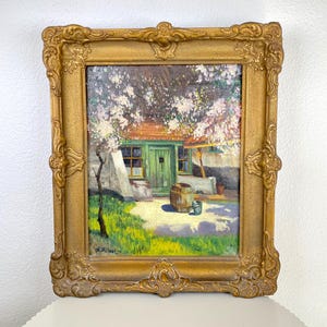 Puede incluir: Pintura al óleo en un marco dorado ornamentado que representa una escena de una cabaña. La cabaña tiene una puerta verde y un tejado rojo, con un árbol floreciente encima. Un barril de madera y un pequeño cubo están en el jardín. Firma del artista visible.
