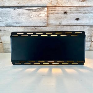 Op de afbeelding: Een rechthoekige, zwarte metalen wandlamp met horizontale sleuven die warm licht uitstralen. De lamp is gemonteerd op een verweerde, lichtgekleurde houten muur. Het licht creëert een schaduwpatroon op het witte oppervlak eronder.