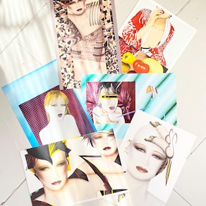 Op de afbeelding: Een verzameling mode-illustratie kunstprints met gestileerde portretten van vrouwen. De prints tonen gedurfde make-up, geometrische patronen en levendige kleuren, met een retro esthetiek. Het kunstwerk is gerangschikt op een wit houten oppervlak.