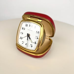 May include: A vintage Junghans travel alarm clock with a square white face, black Arabic numerals, and black hands. The clock is housed in a red, hinged, clam-shell style case with a gold-toned metal trim around the clock face and case edge.