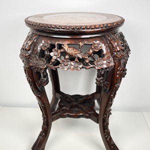 May include: A dark brown wooden pedestal table with a round top featuring a central stone inlay. The apron and legs are intricately carved with floral motifs and bird figures, supported by a cross-shaped stretcher base with detailed carvings.