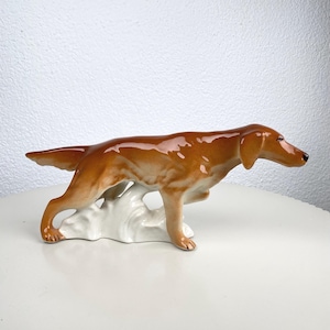 Op de afbeelding: Een keramieken beeldje van een jachthond, met een glanzende bruine vacht en een witte basis. De hond is gedetailleerd gebeeldhouwd en vangt een moment van alertheid. Een decoratief item voor in huis, ongeveer 20 cm lang.