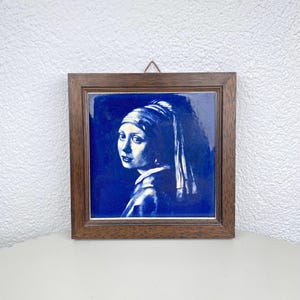 Op de afbeelding: Een ingelijste keramische tegel met een blauw en wit portret van een vrouw met een parel oorbellen. Het kunstwerk is geplaatst in een donkerbruine houten lijst, wat een klassieke en elegante uitstraling creëert. De afbeelding is een reproductie van een beroemd schilderij.