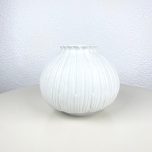 May include: A white ceramic vase with a textured, vertical ribbed design. The vase has a round body and a ruffled top. The vase is sitting on a light-colored surface.