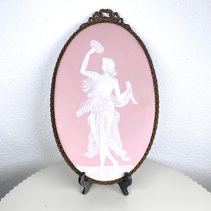 Puede incluir: Cameo ovalado rosa con una figura clásica blanca sosteniendo un plato, enmarcado en un marco color bronce. El marco tiene un borde de cuentas y una parte superior decorativa. El cameo se exhibe sobre un soporte negro.