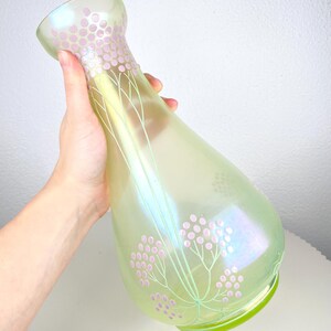 May include: Iridescent green glass vase with a flared top and a rounded base. The vase is decorated with pink floral patterns and delicate green stems. The vase has a pearlescent sheen.