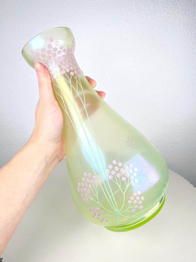May include: Iridescent green glass vase with a flared top and a rounded base. The vase is decorated with pink floral patterns and delicate green stems. The vase has a pearlescent sheen.