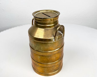 May include: A vintage brass money box in the shape of a milk churn. It features a slot for coins on the top and two small handles. The body is banded horizontally. The brass has a warm, aged patina.