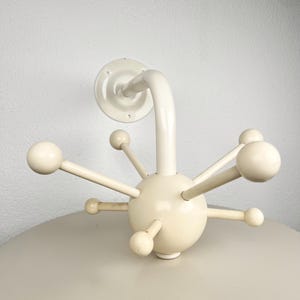 Pode incluir: Um cabide de parede na cor creme com uma esfera central e múltiplos braços radiantes, cada um terminando em um gancho liso em forma de bola. A estrutura inclui um braço de suporte curvo fixado a uma placa de montagem circular com furos.