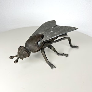 Op de afbeelding: Een bronzen metalen sculptuur van een vlieg. Het insect heeft gedetailleerde vleugels, poten en antennes. De sculptuur staat op een wit oppervlak en toont de ingewikkelde details van het kunstwerk.