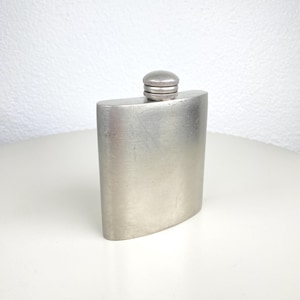 Op de afbeelding: Een zilverkleurige heupfles met een afgeronde rechthoekige vorm en een schroefdop. De fles heeft een geborstelde metalen afwerking. De dop is donkerder zilver en heeft een geribbelde textuur voor grip. De fles is ongeveer 12 cm hoog.