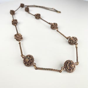 Puede incluir: Un collar vintage de bronce con colgantes circulares de filigrana unidos por finas cadenas. El collar tiene un aspecto antiguo, con diseños florales detallados en cada colgante. El collar se muestra sobre una superficie blanca. El collar mide aproximadamente 45 cm de largo.
