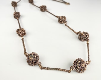 Puede incluir: Un collar vintage de bronce con colgantes circulares de filigrana unidos por finas cadenas. El collar tiene un aspecto antiguo, con diseños florales detallados en cada colgante. El collar se muestra sobre una superficie blanca. El collar mide aproximadamente 45 cm de largo.