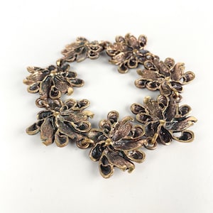 Op de afbeelding: Een vintage bronzen armband met een bloemenontwerp. De armband bestaat uit bloemvormige schakels, elk met gedetailleerde bloembladtexturen en een donkere, verouderde afwerking. De schakels zijn verbonden door kleine, cirkelvormige connectoren.