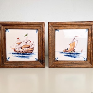 Op de afbeelding: Twee vierkante keramische tegels in donkerhouten lijsten. Elke tegel toont een handgeschilderde illustratie van een zeilschip op blauw water. De hoeken van de tegels zijn versierd met kleine blauwe bloemmotieven op een witte achtergrond.