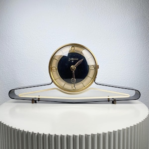 May include: A vintage clock with a gold-tone bezel and a black face. The clock is set on a clear, angled stand with a black frame. The clock's hands are black, and the numbers are gold. The clock is displayed on a white surface.