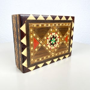 Puede incluir: Una caja rectangular de madera con intrincados patrones geométricos incrustados. La caja presenta un exterior marrón oscuro con una tapa decorativa que muestra un diseño central en forma de estrella en colores rojo, verde y crema. Los lados tienen un patrón repetitivo.