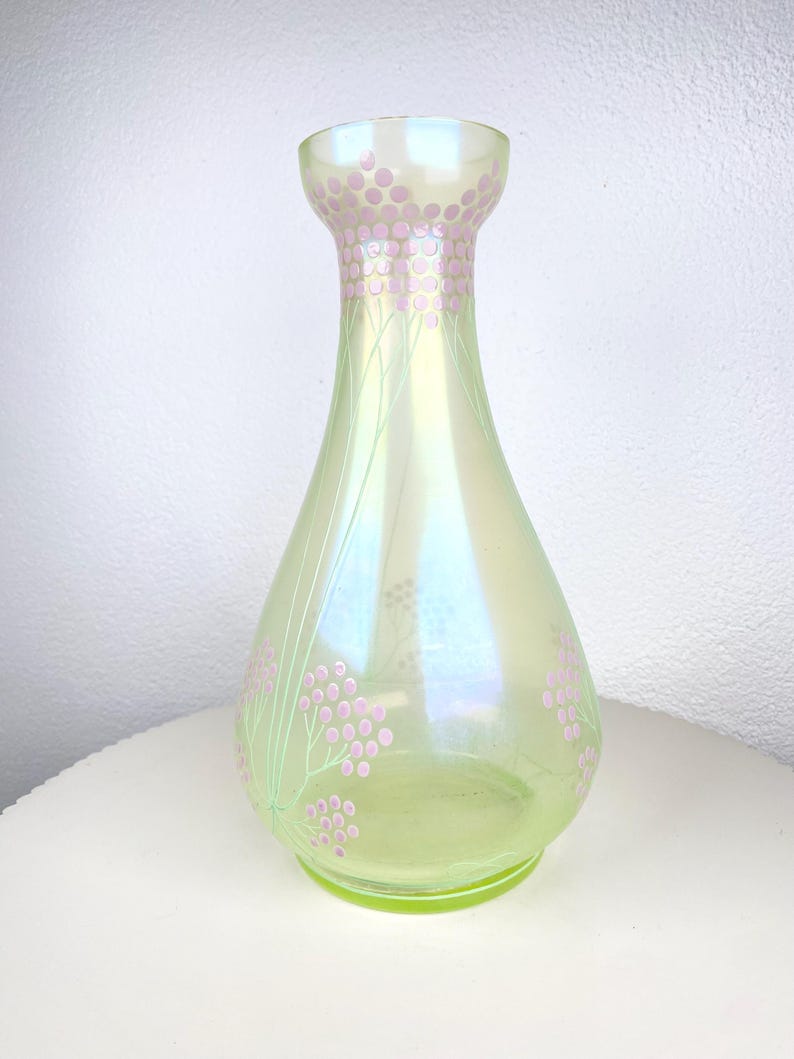 May include: A pearlescent green glass vase with a flared neck and a rounded body. The vase is decorated with pink floral patterns and delicate green stems. The vase is approximately 23 cm tall.