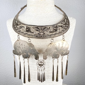 Op de afbeelding: Zilverkleurige statement ketting met een sierlijke kraag met gegraveerde patronen. Twee olifantenhangers hangen eronder, verbonden door een bloemvormig middelpunt, met bungelende kegelvormige bedels. De ketting wordt getoond op een witte mannequin.