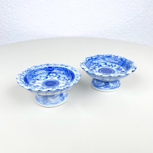 Op de afbeelding: Twee bijpassende blauw-witte porseleinen schalen op voet. De schalen hebben een geschulpte rand en een gedetailleerd bloemen- en geometrisch patroon. Ze staan op een korte, versierde voet. De schalen staan op een wit oppervlak.