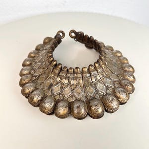 Könnte beinhalten: Ein breites, starres Metall-Halsband, bestehend aus einer Reihe verbundener, geschwungener Segmente. Jedes Segment ist mit einem geprägten Rautenmuster und einer Reihe abgerundeter, texturierter Noppen am Außenrand verziert, in einem angelaufenen Silberton.