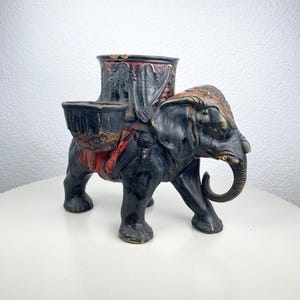 Puede incluir: Figura de elefante negro y rojo con un recipiente decorativo en su espalda. El elefante tiene una trompa y colmillos detallados, con detalles dorados en la cabeza y las orejas. El recipiente tiene un interior rojo y está diseñado para guardar objetos.