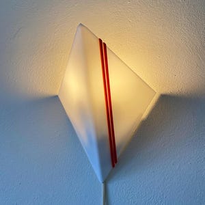 May include: A white diamond-shaped wall sconce with two red stripes running diagonally across the front. The sconce is lit and casting a warm glow.