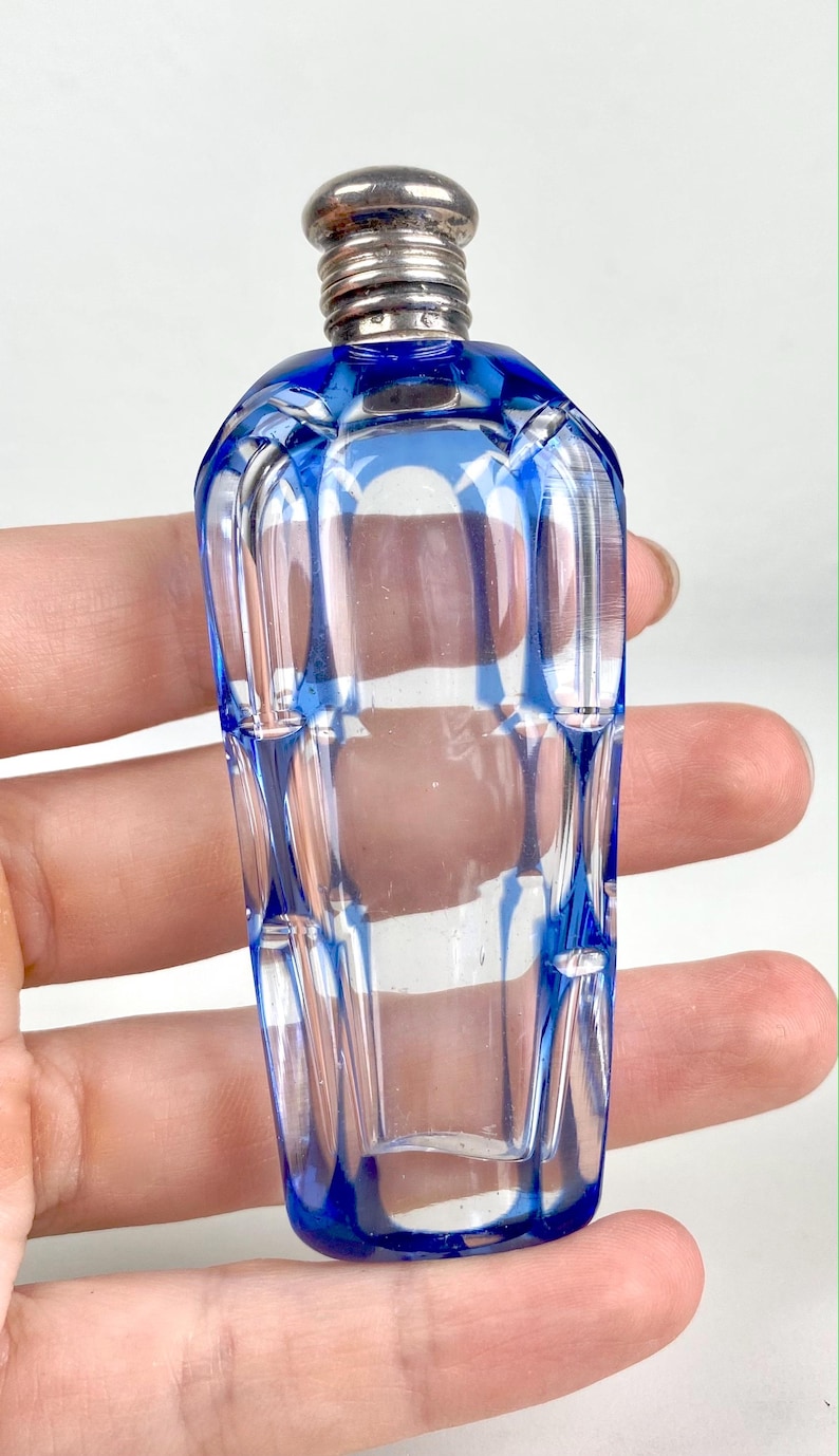 May include: A small, clear glass perfume bottle with a blue geometric design. The bottle has a silver-colored metal cap. The bottle is held in a hand.