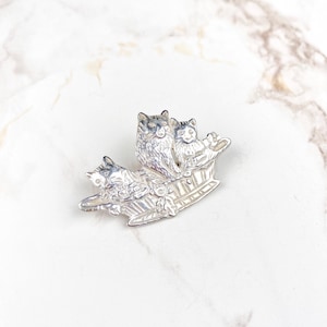 Op de afbeelding: Een zilverkleurige broche met drie gestileerde katten in een bootvormig ontwerp. De katten hebben gedetailleerde vacht en zijn zo gerangschikt dat ze naar voren kijken. De broche staat tegen een witte marmeren achtergrond.
