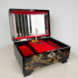 May include: A black lacquer music box featuring gold landscape illustrations on the exterior, a red velvet interior lining, a rectangular mirror inside the lid, and a small ballerina figurine that stands on a central platform when the lid is open.
