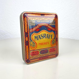 Könnte beinhalten: Vintage Masraff Frères ägyptische Zigaretten-Blechdose. Die rechteckige Dose hat ein gold-, blau- und rotes Design mit Text, darunter "Masraff Frères" und "Cairo". Ein Sammlerstück.