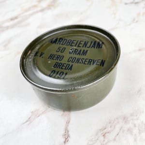 Puede incluir: Una lata de metal verde oliva con el texto "AARDBEIENJAM 50 GRAM V. HERO CONSERVEN BREDA 0191" impreso en negro. La lata es redonda y tiene una tapa ligeramente abovedada. El fondo es un patrón de mármol.