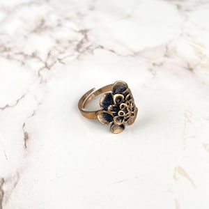 May include: A bronze-colored ring with a floral design. The ring features a cluster of petals and a band that adjusts to fit different finger sizes. The ring is set against a white marble background.
