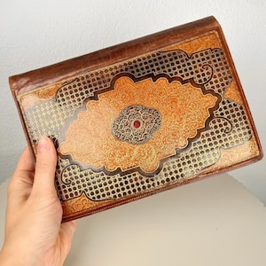 May include: A rectangular brown leather portfolio featuring an intricate embossed pattern with gold-toned detailing. The centre displays a decorative medallion with a small red circular accent, surrounded by ornate filigree and geometric grid motifs.