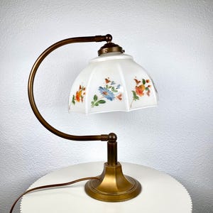 Puede incluir: Lámpara de escritorio de estilo vintage con brazo y base curvados de color bronce. La pantalla es blanca con un estampado floral en naranja, azul y verde. La lámpara está diseñada para colocarse sobre un escritorio o mesa.