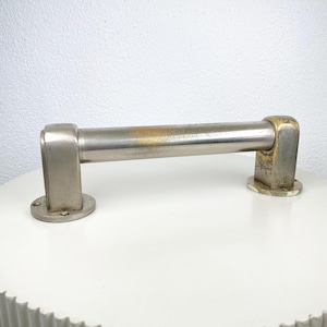May include: A silver-toned metal handle with a cylindrical bar and rectangular end pieces. The handle shows signs of age with some discoloration. The ends have circular mounting plates. The handle is likely for a cabinet or drawer.