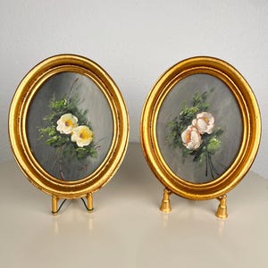 Op de afbeelding: Een paar ovale schilderijen in sierlijke gouden lijsten, elk tentoongesteld op een kleine gouden standaard. De schilderijen tonen bloemstukken in olieverfstijl met witte en gele bloesems tegen een gedempte grijze achtergrond, als een set.