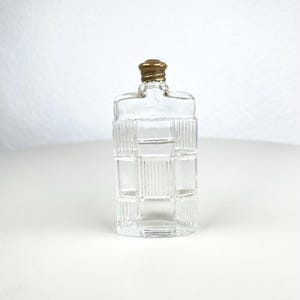 Op de afbeelding: Een heldere glazen fles met een rechthoekige vorm en een goudkleurige dop. De fles heeft een gestructureerd, rasterachtig patroon aan de voor- en zijkanten. De fles is leeg en staat op een wit oppervlak.