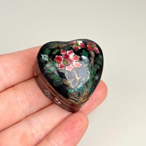 Op de afbeelding: Een hartvormig doosje met een zwarte emaille basis en een kleurrijk bloemendesign in tinten roze, groen en wit. Het deksel toont een gedetailleerd bloemmotief, terwijl de zijkanten van het doosje zijn versierd met kleinere florale accenten.