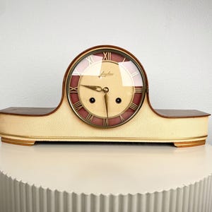 May include: A mantel clock with a low, elongated wooden base featuring a cream-colored textured front. The circular clock face has a cream center, a dark red outer ring with gold Roman numerals, gold hands, and the Junghans brand name in script.