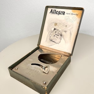 Op de afbeelding: Een olijfgroene metalen Allegro mod. Special scheermesjesslijper in een open doos. De binnenzijde van het deksel bevat een gedrukte illustratie van handen die het apparaat gebruiken, terwijl de basis het mechanische slijpmechanisme bevat.