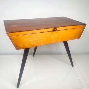 Op de afbeelding: Een vintage houten naaikist met een donkerbruin deksel en een lichtbruine behuizing. De kist heeft een kleine, ronde houten knop en wordt ondersteund door vier donkerbruine, schuine poten. Vintage stijl.