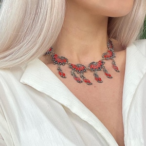 Op de afbeelding: Een zilverkleurige metalen ketting met een reeks waaiervormige schakels gevuld met felrood emaille. Het ontwerp bevat meerdere bungelende elementen die aan de hoofdketting hangen, wat zorgt voor een gelaagd effect rond de hals.