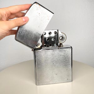 Op de afbeelding: Een zilverkleurige metalen aansteker met een geborstelde afwerking en een rechthoekige behuizing. Het deksel staat open en toont de interne schoorsteen met ronde ventilatiegaten en een gekarteld wieltje. Het metaal vertoont lichte gebruikssporen.