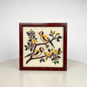 Op de afbeelding: Een vierkant kruissteek borduurwerk met twee gele en zwarte vogels op bruine takken met groene bladeren en kleine paarse en gele bloemen, geplaatst in een vierkante lijst met een donkerroodbruine houtnerfstructuur.